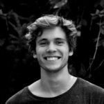 A black and white portrait of a young man smiling outdoors with natural background.